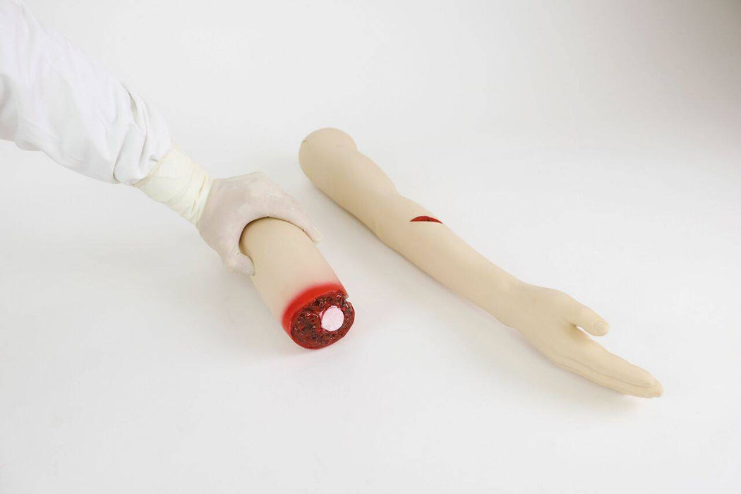 A gloved hand holds a severed prosthetic arm; a second prosthetic arm with a simulated wound lies nearby on a white surface.