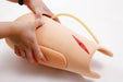 A training arm model is being manipulated; a tube is inserted near a simulated wound. The setting appears to be a medical training facility.