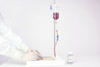 A blood bag hangs from a stand, undergoing a transfusion process; a gloved hand is near the tubing, in a sterile medical setting.