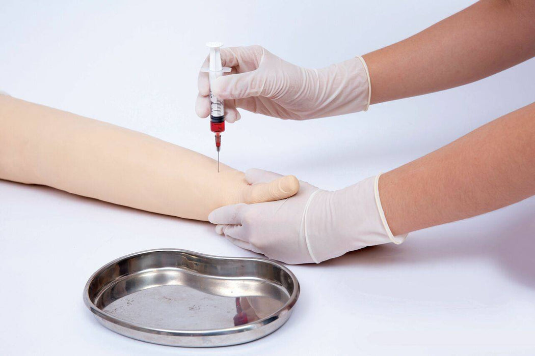 A gloved hand injects a simulated arm with a syringe containing red liquid on a white surface near a kidney-shaped metal tray.