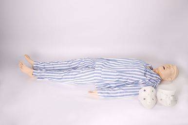 A medical mannequin lies supine on a white surface; it wears striped pajamas and has neck braces nearby.