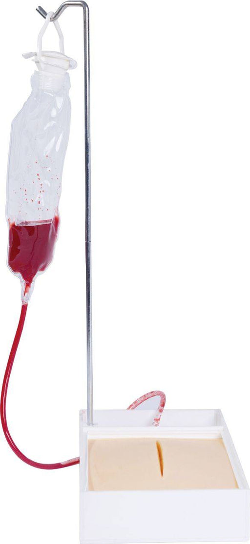 A blood bag hangs from a stand, its tube connected to a simulated incision on a training pad. The setup is likely for medical training.