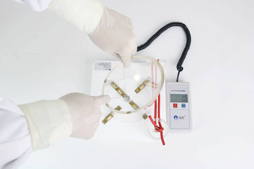 Gloved hands manipulate a transparent, circular device with internal components. A digital scale displays “0”. The setting is a clean, white laboratory.
