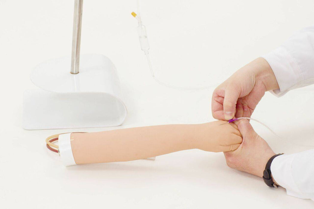 A training arm is having an IV inserted by hands in white coats, on a white surface; a stand supports the arm.