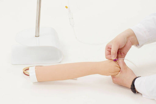 A training arm is having an IV inserted by hands in white coats, on a white surface; a stand supports the arm.