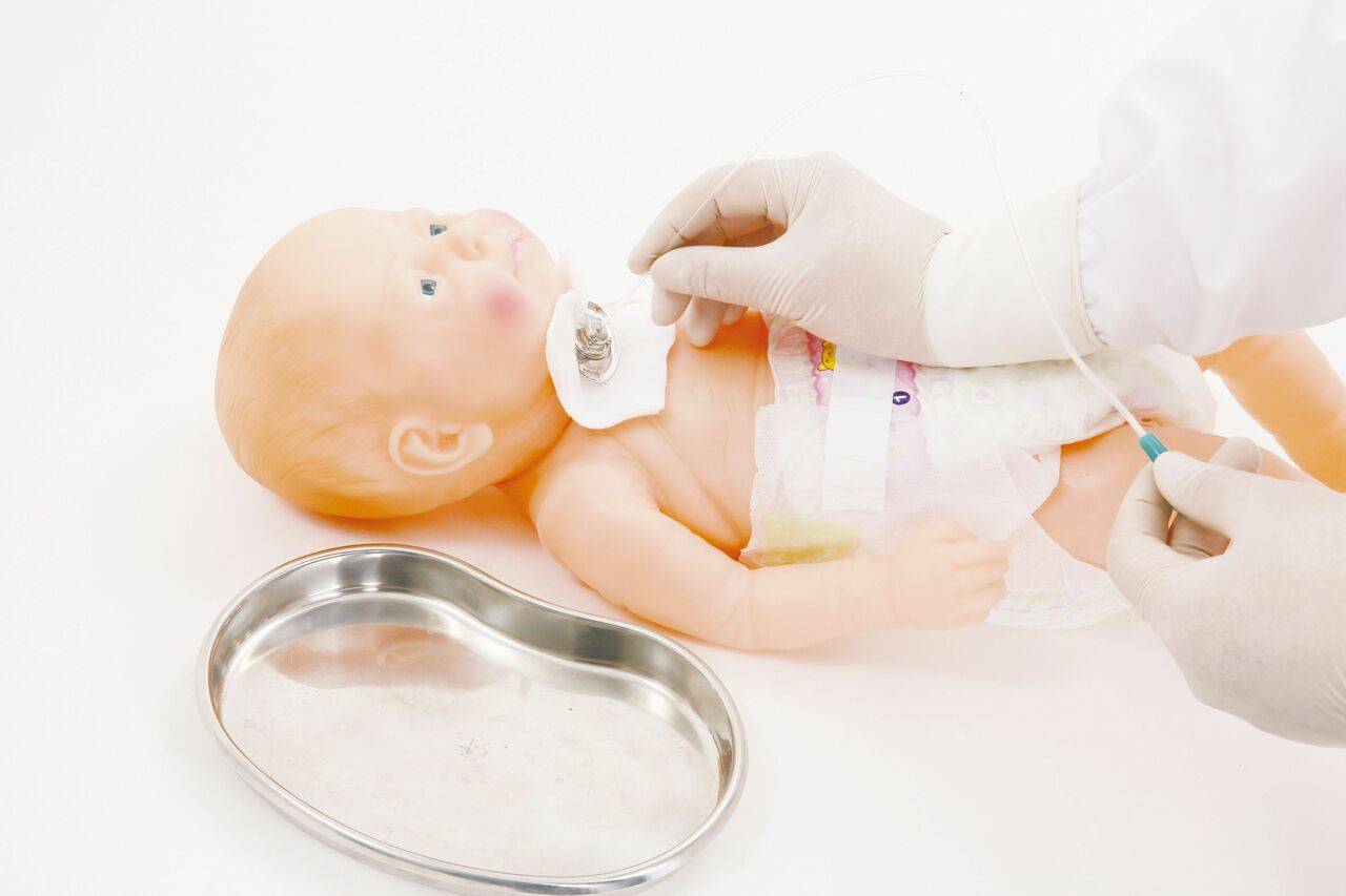 A baby doll receives a medical procedure; gloved hands manipulate a tube near its abdomen; a kidney-shaped tray is on a white surface.