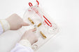 Gloved hands manipulate a small object with a needle in a clear, plastic container with attached golden components and red tubing, set against a white backdrop.