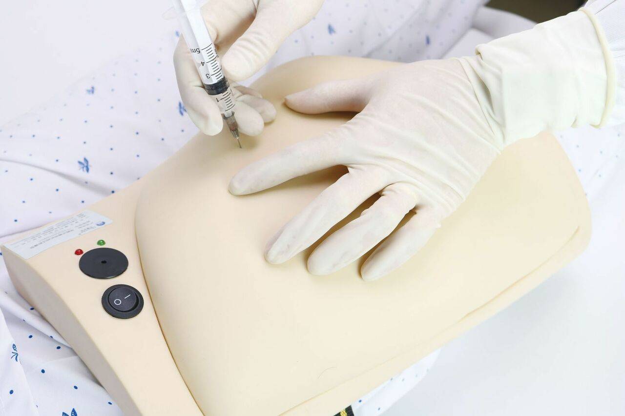 Gloved hands inject a training model; the model is a pale, flesh-toned torso, lying on a bed.