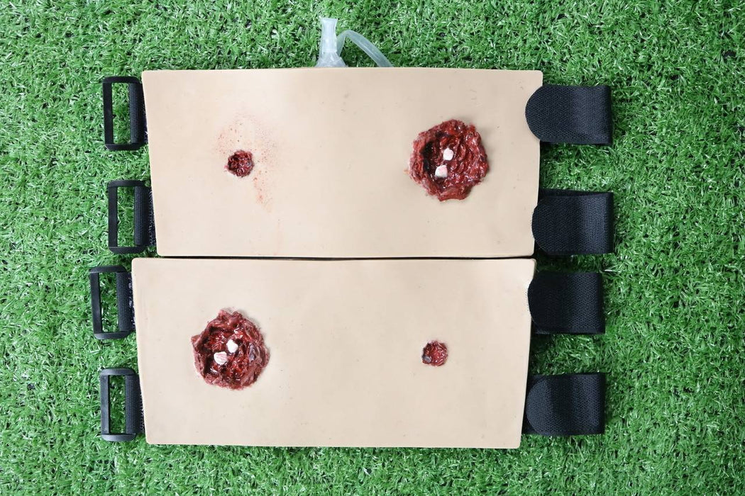 Two tan, padded training aids, simulating gunshot wounds, lie on artificial turf. Each aid has two simulated wounds; one larger and one smaller. They are fastened with black straps.