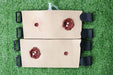 Two tan, padded training aids, simulating gunshot wounds, lie on artificial turf. Each aid has two simulated wounds; one larger and one smaller. They are fastened with black straps.