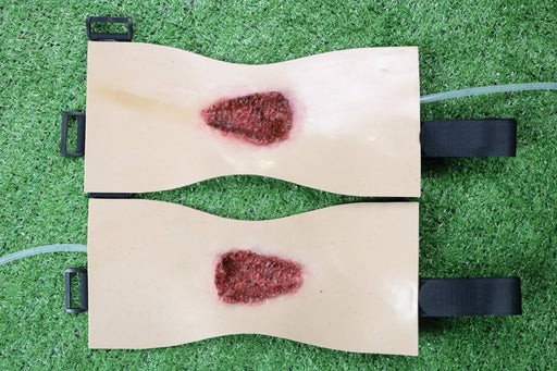 Two tan fabric pads, each with a simulated wound, are fastened with black straps. They lie on a green surface.
