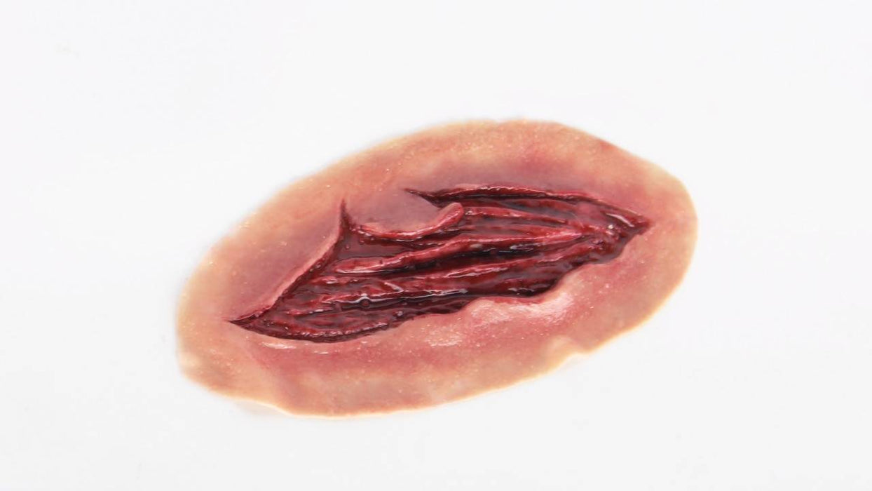 A flesh-colored, oval prosthetic sits on a white surface; a gash filled with dark red, fleshy-looking material forms a central wound.