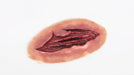 A flesh-colored, oval prosthetic sits on a white surface; a gash filled with dark red, fleshy-looking material forms a central wound.