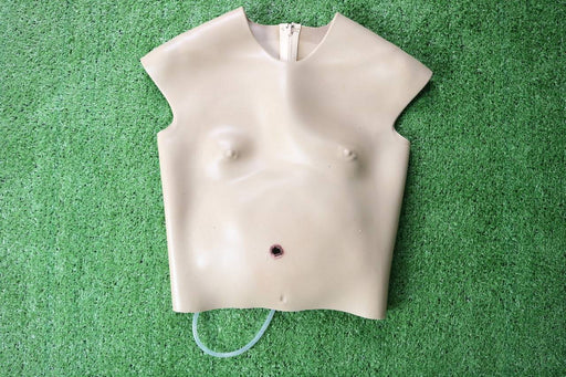 A tan, sleeveless torso mannequin with a simulated wound rests on artificial turf. A clear tube extends from its lower abdomen.