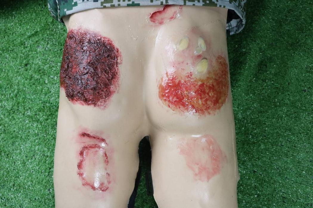A training dummy displays simulated burns and lacerations on its buttocks and thighs; it rests on artificial turf.