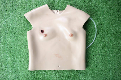 A tan, sleeveless torso mannequin rests on green turf. It simulates wounds and has attached tubing.