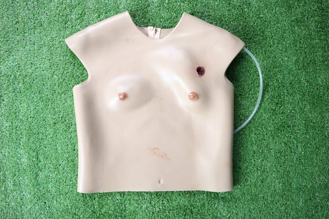 A beige torso mannequin with simulated gunshot wound and nipples rests on artificial turf. A clear tube is looped nearby.
