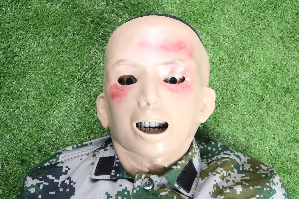 A training dummy, depicting facial injuries, lies on artificial turf. The dummy is wearing a camouflage uniform.
