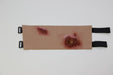 A tan, rectangular training aid depicts two simulated wounds; it features black straps at each end.
