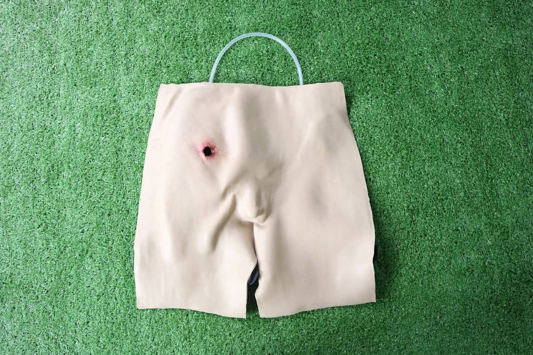 A tan, cloth torso simulator displays a simulated gunshot wound; a clear tube is looped over its top. It rests on artificial turf.