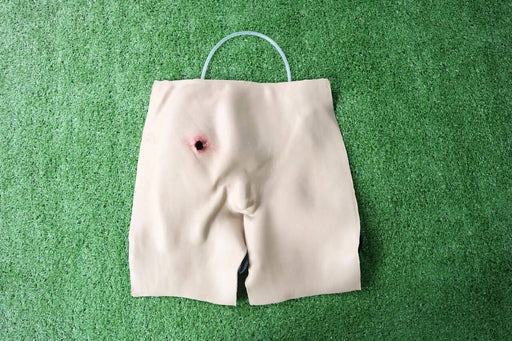 A tan, cloth torso simulator displays a simulated gunshot wound; a clear tube is looped over its top. It rests on artificial turf.