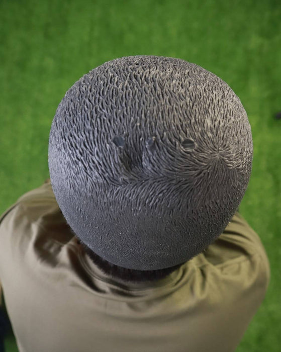 A dark, textured head rests atop a person wearing an olive-green shirt. The person is situated on a green surface.