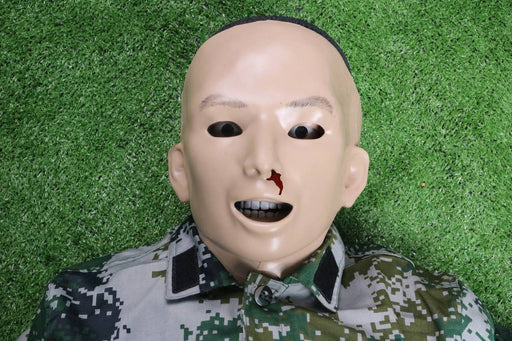 A training dummy lies on artificial turf; it wears a camouflage uniform and has simulated blood on its nose.