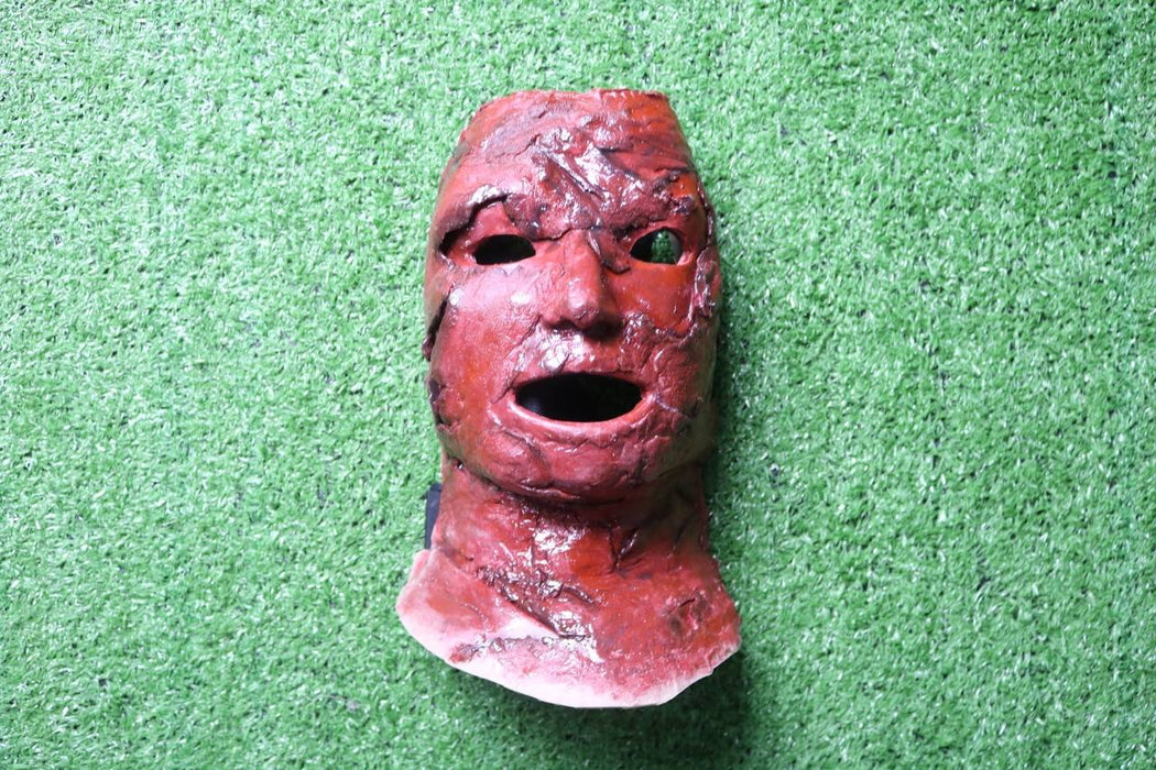 A red, textured mask, resembling burned flesh, rests on artificial turf.