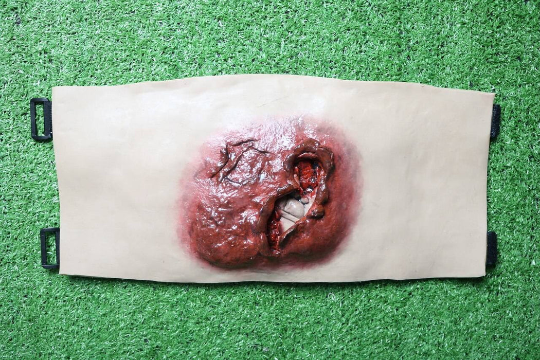 A realistic, simulated wound is affixed to a tan backing with black straps; it rests on artificial turf.