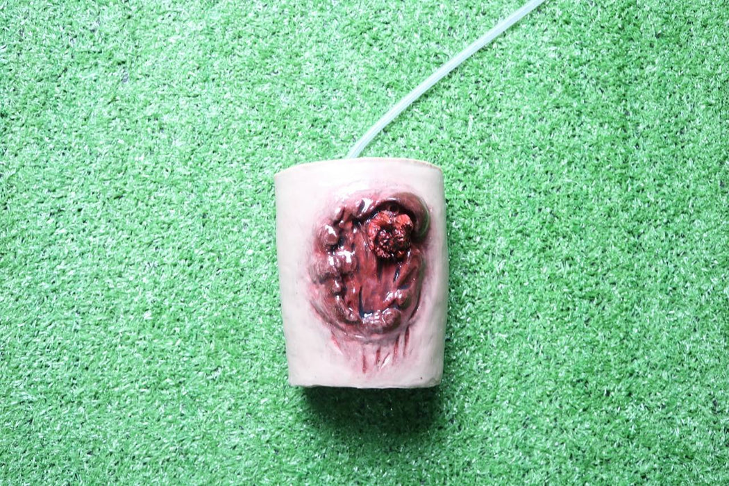 A gruesome wound replica rests on artificial turf; a clear tube extends from it.