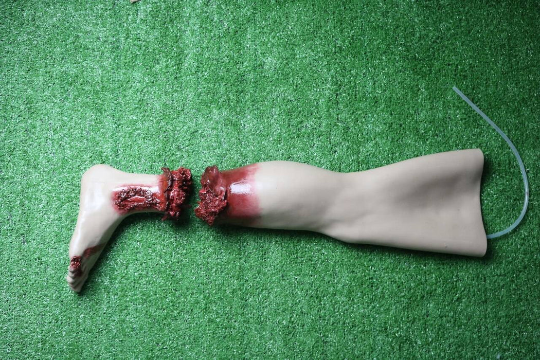 A severed prosthetic leg lies on artificial turf. The leg is broken into two parts, with simulated blood around the break. A clear tube extends from the lower section.