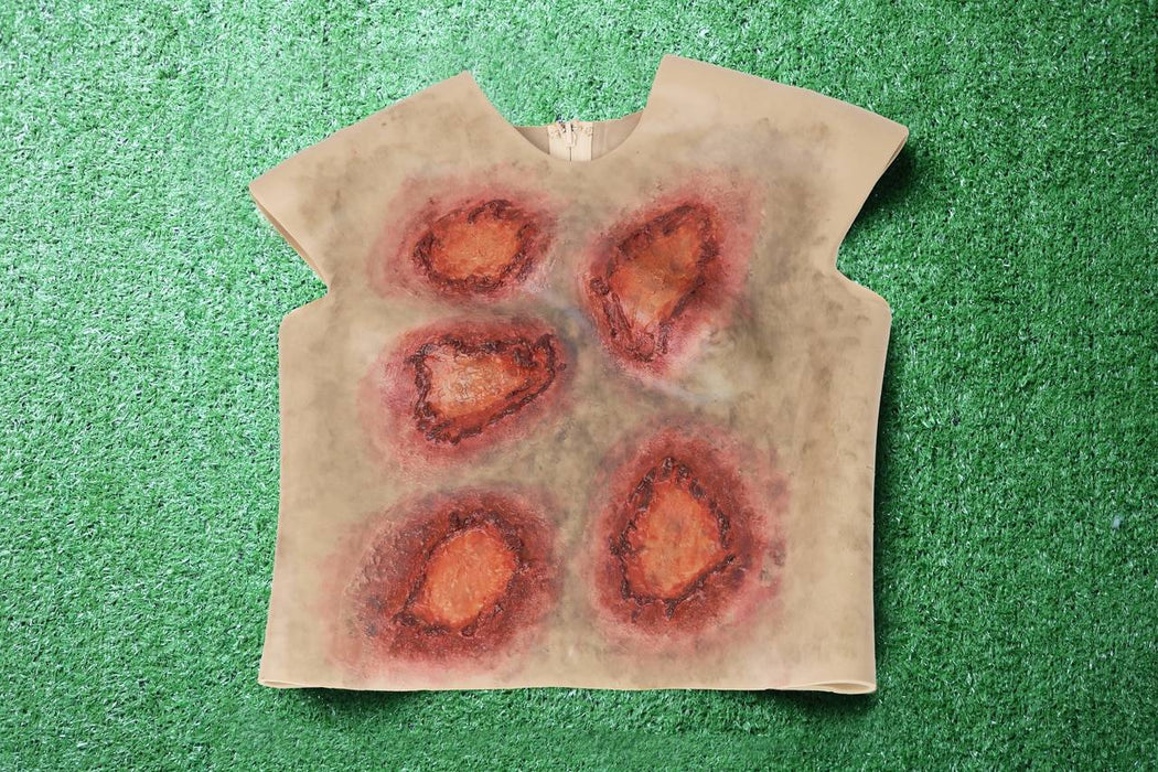 A sleeveless shirt depicting simulated wounds rests on artificial turf. The wounds are painted in shades of red and brown, suggesting injury.
