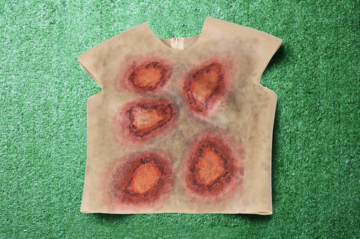 A sleeveless shirt depicting simulated wounds rests on artificial turf. The wounds are painted in shades of red and brown, suggesting injury.