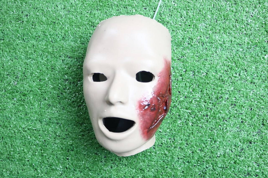 A realistic face mask, displaying a bloody injury, rests on artificial turf.