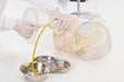 Gloved hands use tools on a plastic training dummy within a clear container, with a yellow tube draining fluid into metal containers on a white surface.