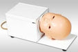 A baby mannequin head rests inside a white box-like device on a white surface; the device appears to be for medical training or practice.