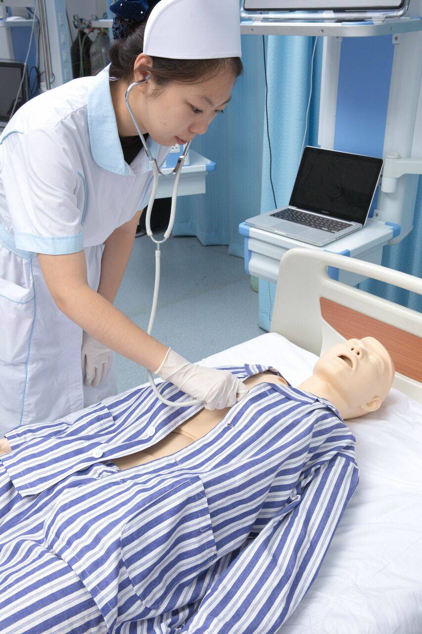 A nurse uses a stethoscope to examine a medical training mannequin lying on a hospital bed in a clinical setting.