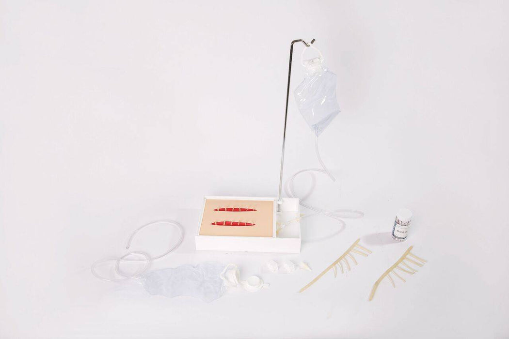 A medical training kit displays a simulated skin pad with two deep, stitched lacerations. The kit includes an IV bag on a stand, tubing, sutures, gauze, and a medicine bottle, all presented against a stark white background.