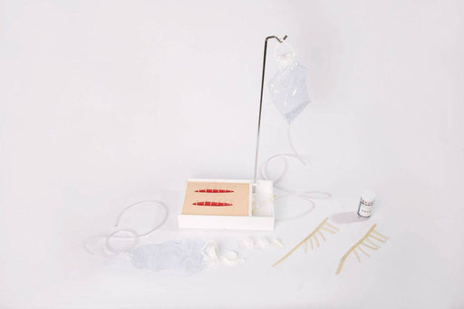 A medical training kit displays a simulated skin pad with two deep, stitched lacerations. The kit includes an IV bag on a stand, tubing, sutures, gauze, and a medicine bottle, all presented against a stark white background.