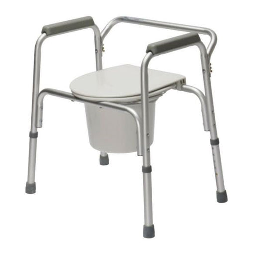 A silver, adjustable commode chair stands, prepared for use, against a plain white background. It has armrests and a container under the toilet seat.