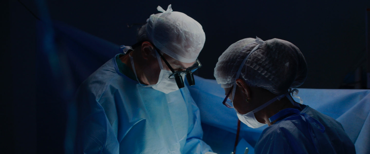 Two surgeons, wearing surgical caps, gowns, masks, and eyewear, perform surgery in a dimly lit operating room.
