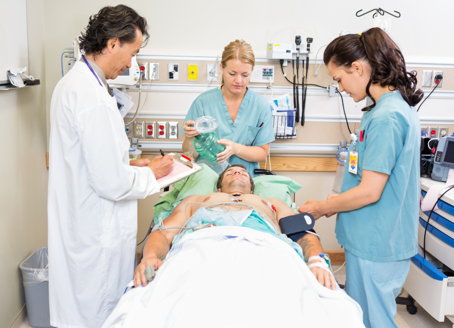 A male patient lies supine on a hospital bed, being monitored and attended to by three medical professionals in scrubs and a lab coat. One is writing notes, another is preparing a mask, and the third is attaching sensors.