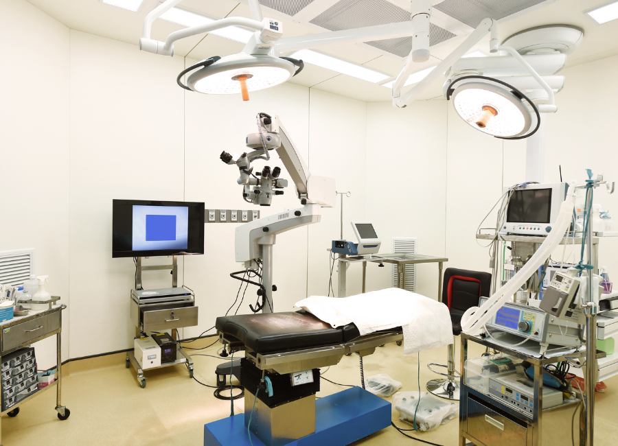 A surgical microscope stands in a modern operating room, surrounded by medical equipment including an operating table and monitors.