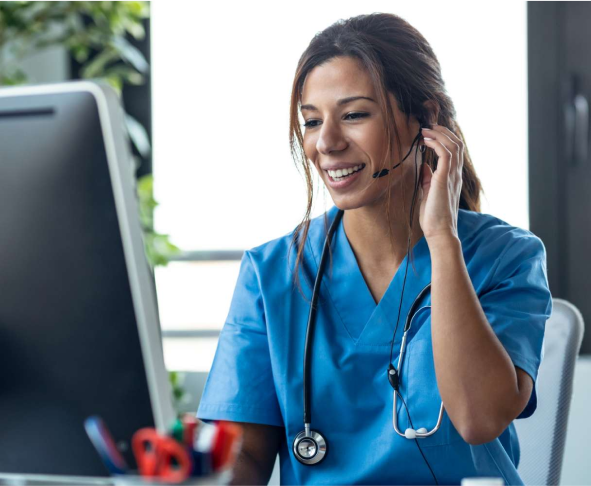 A nurse, wearing scrubs and a headset, uses a computer in a brightly lit office.