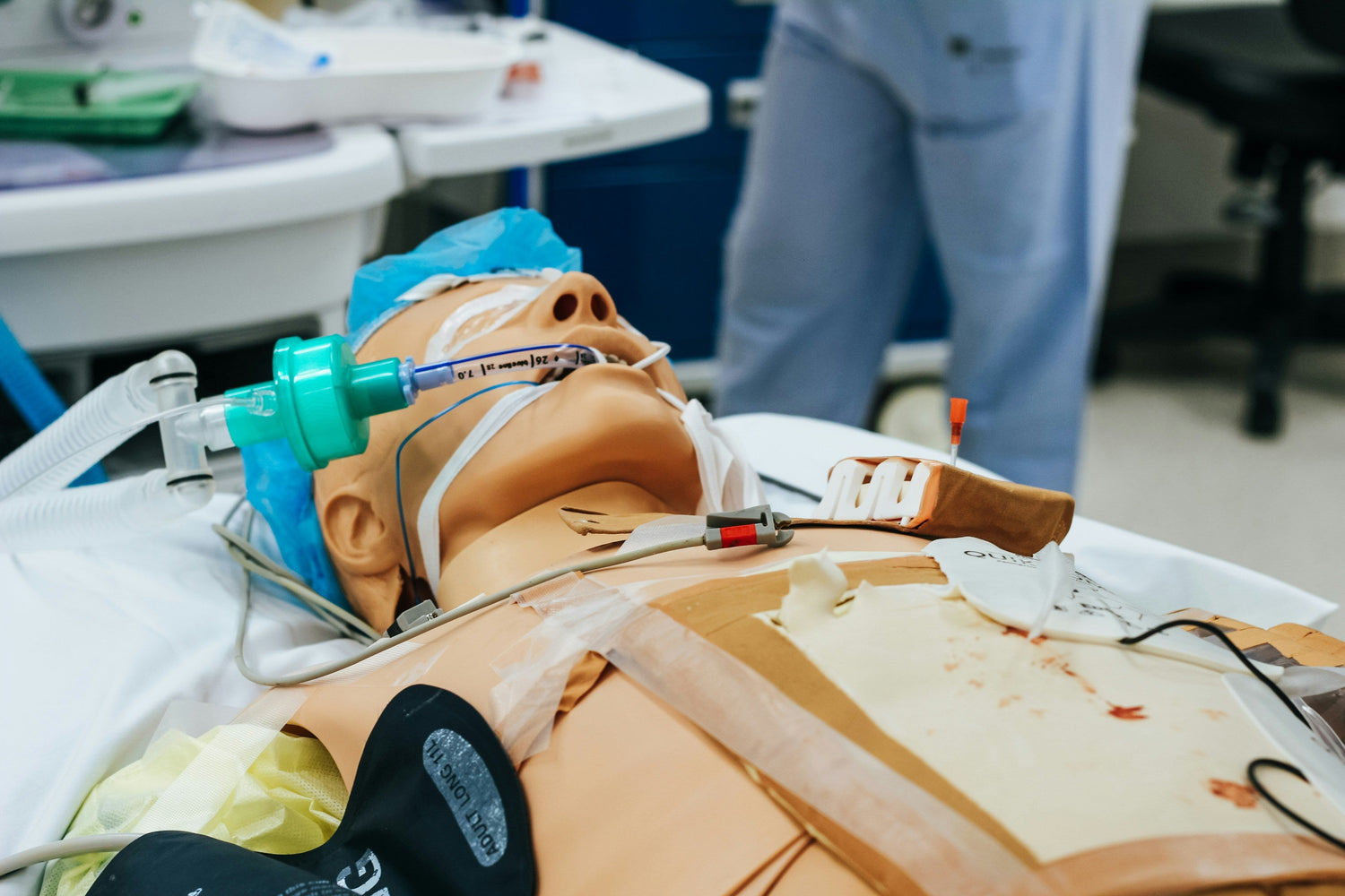 A medical mannequin lies on a table, connected to breathing and monitoring equipment, simulating a patient in a hospital setting. ADULT LONG TH is visible on a black device attached to the mannequin.